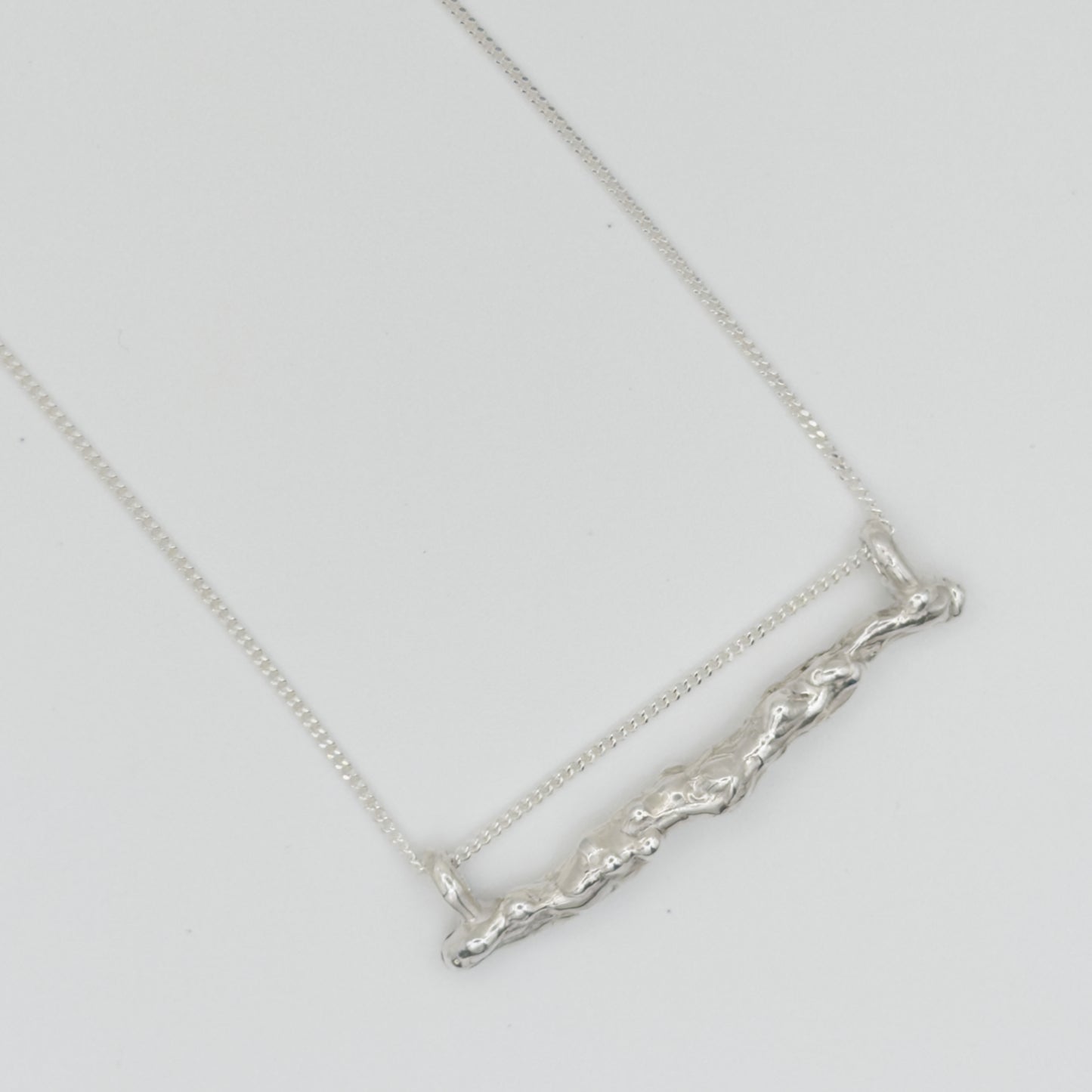 Close up of the Atoll textured silver bar pendant on a chain, pictured on a light plain background.