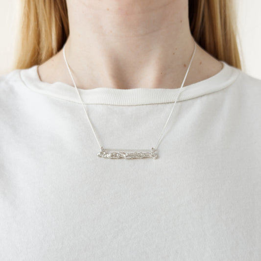 Close up of the Atoll silver necklace, featuring a textured bar pendant. Shown on a person wearing a white top.
