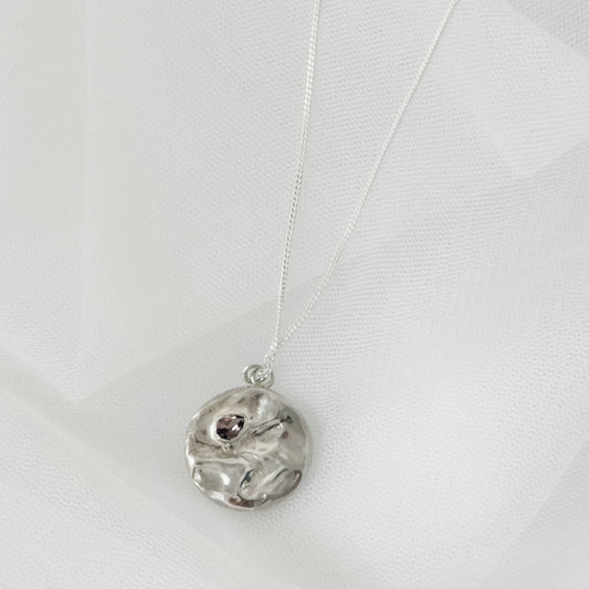 Front view of the Devotion silver necklace, featuring a round pendant. Pictured on a light textured background.