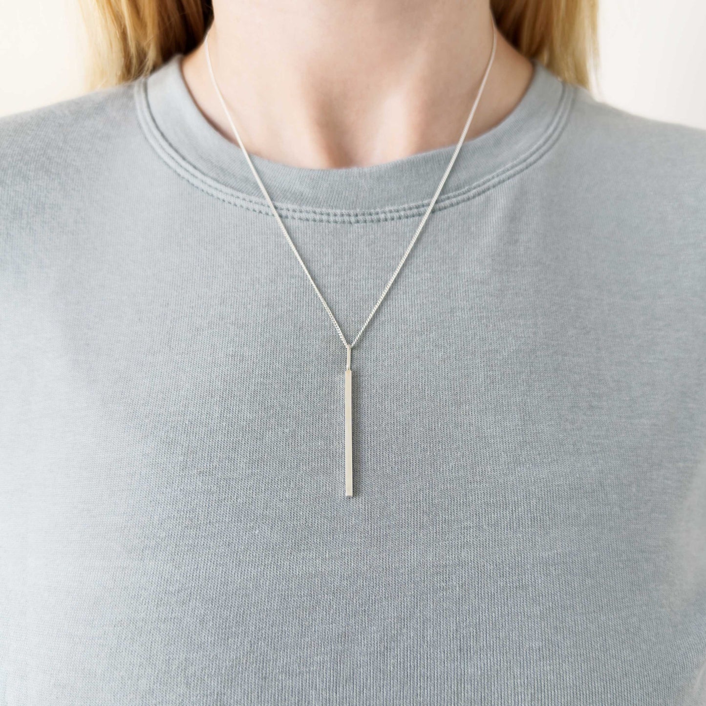 Alignment silver bar pendant on a curb chain. Pictured on a person wearing a light blue-gray top.