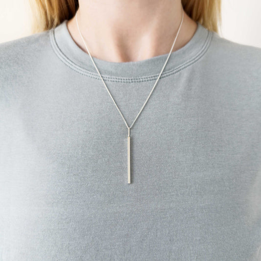 Alignment silver bar pendant on a curb chain. Pictured on a person wearing a light blue-gray top.