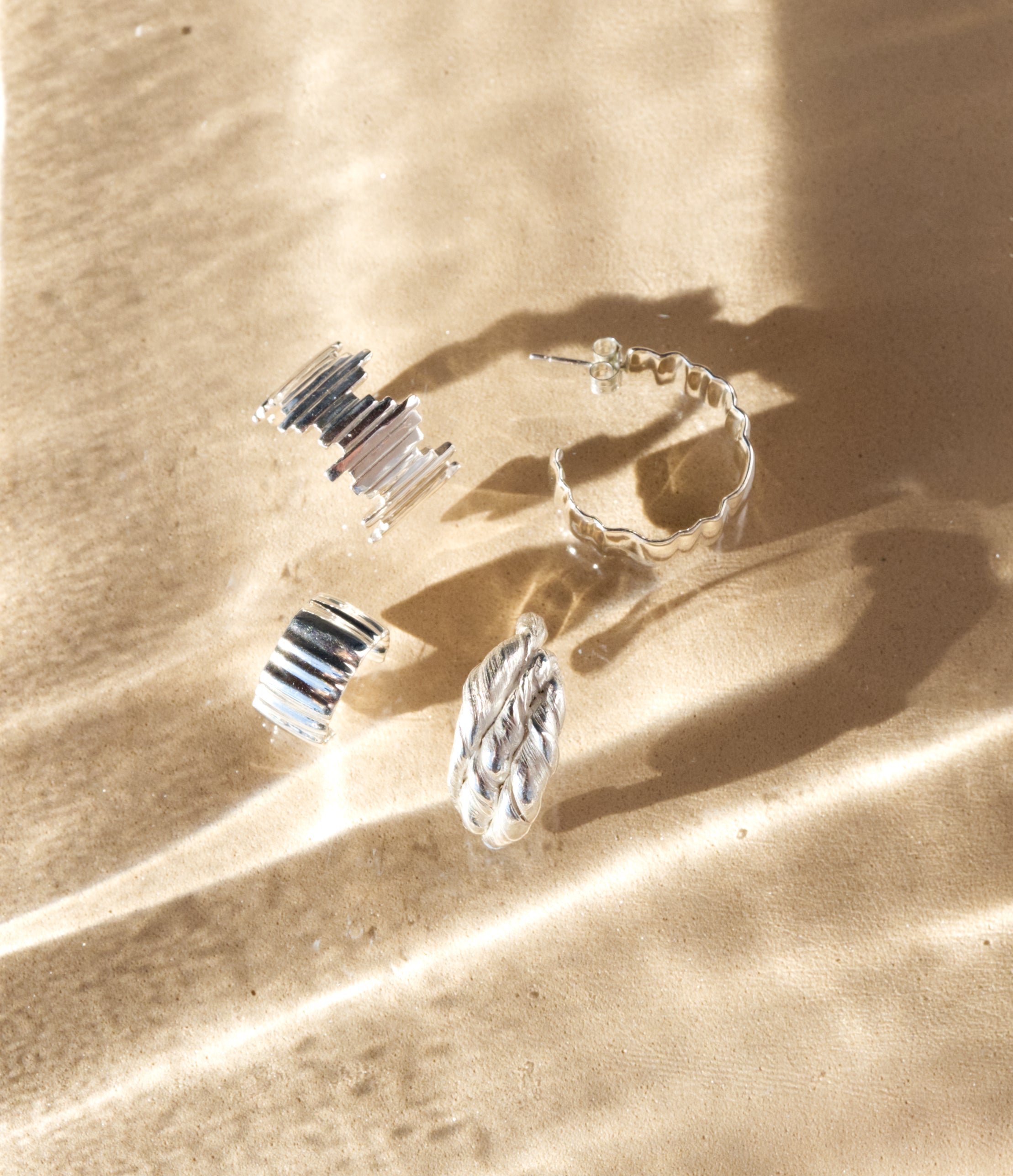 Four different silver hoop earrings: Guidance, Wave Large, Wave Small, and Anchor, pictured on a sandy textured sunlit background.