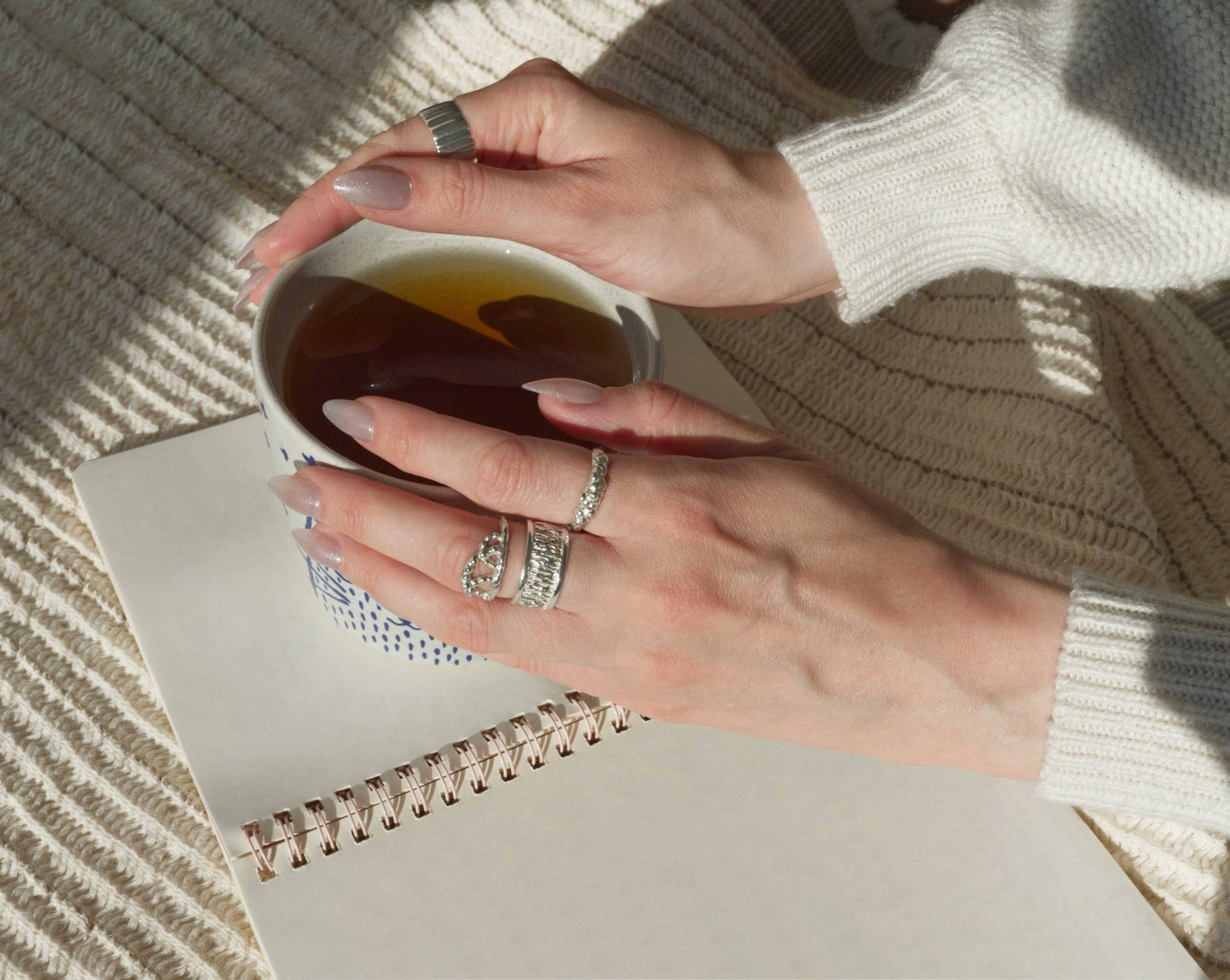 Two hands holding a ceramic mug, wearing stacked sterling silver textural rings, resting over an open notebook in morning light.