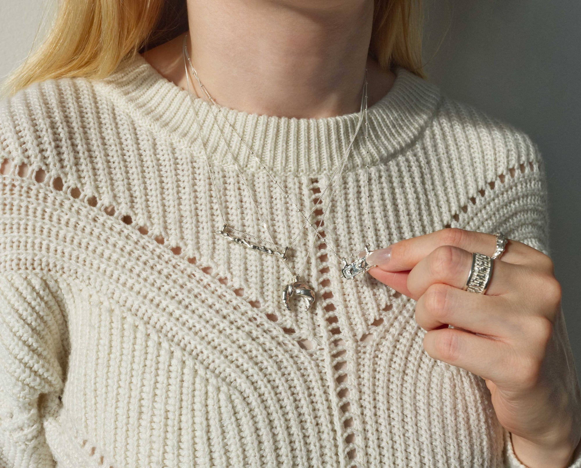 A hand touching a layered sterling silver necklace against a cream knit sweater, with a stacked textural ring visible on the finger, in warm natural light.
