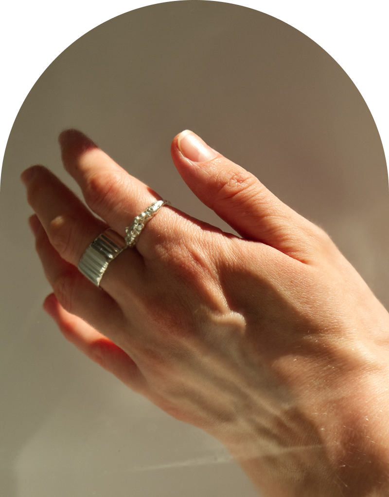A hand in warm natural light wearing two stacked sterling silver rings - a wide ribbed band and a delicate textured ring - against a soft neutral background with an arch crop.