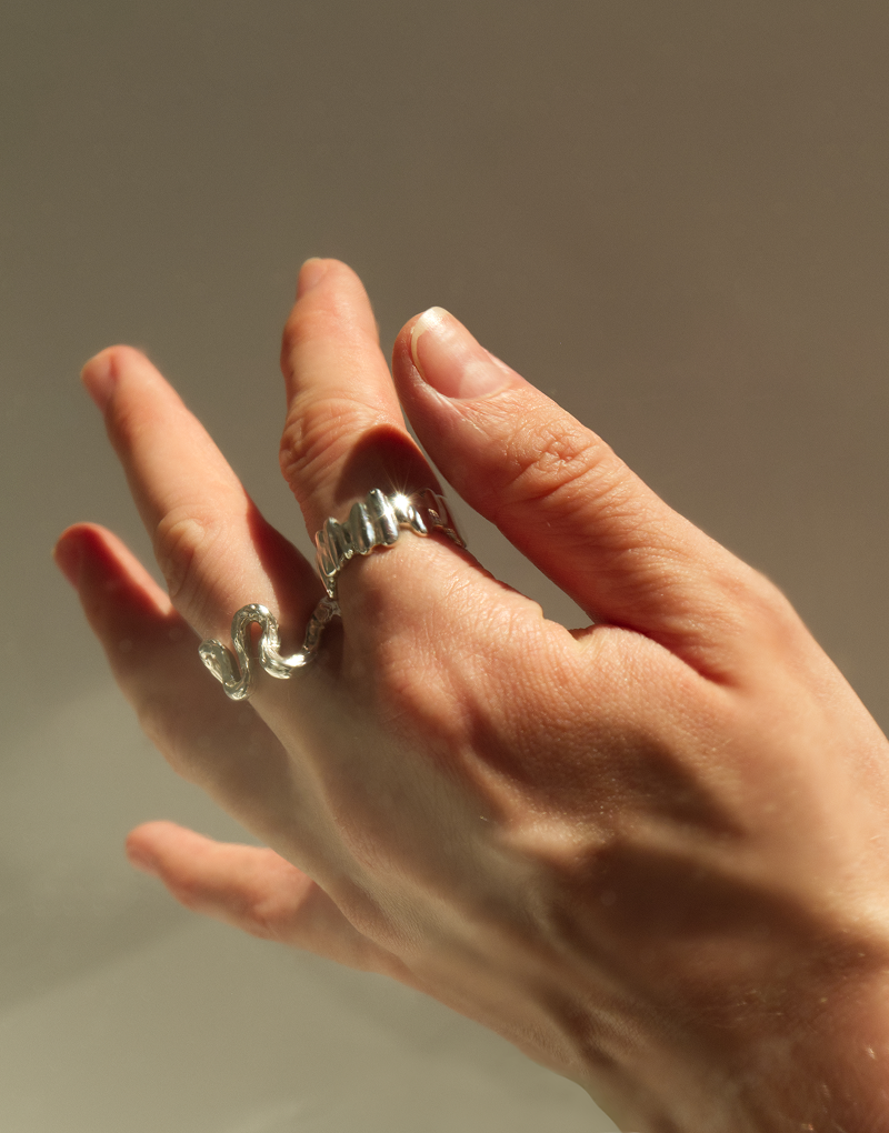 A hand in directional natural light wearing two sterling silver rings - a sculptural wave-form ring on the index finger and a ridged band on the middle finger - against a warm grey background.