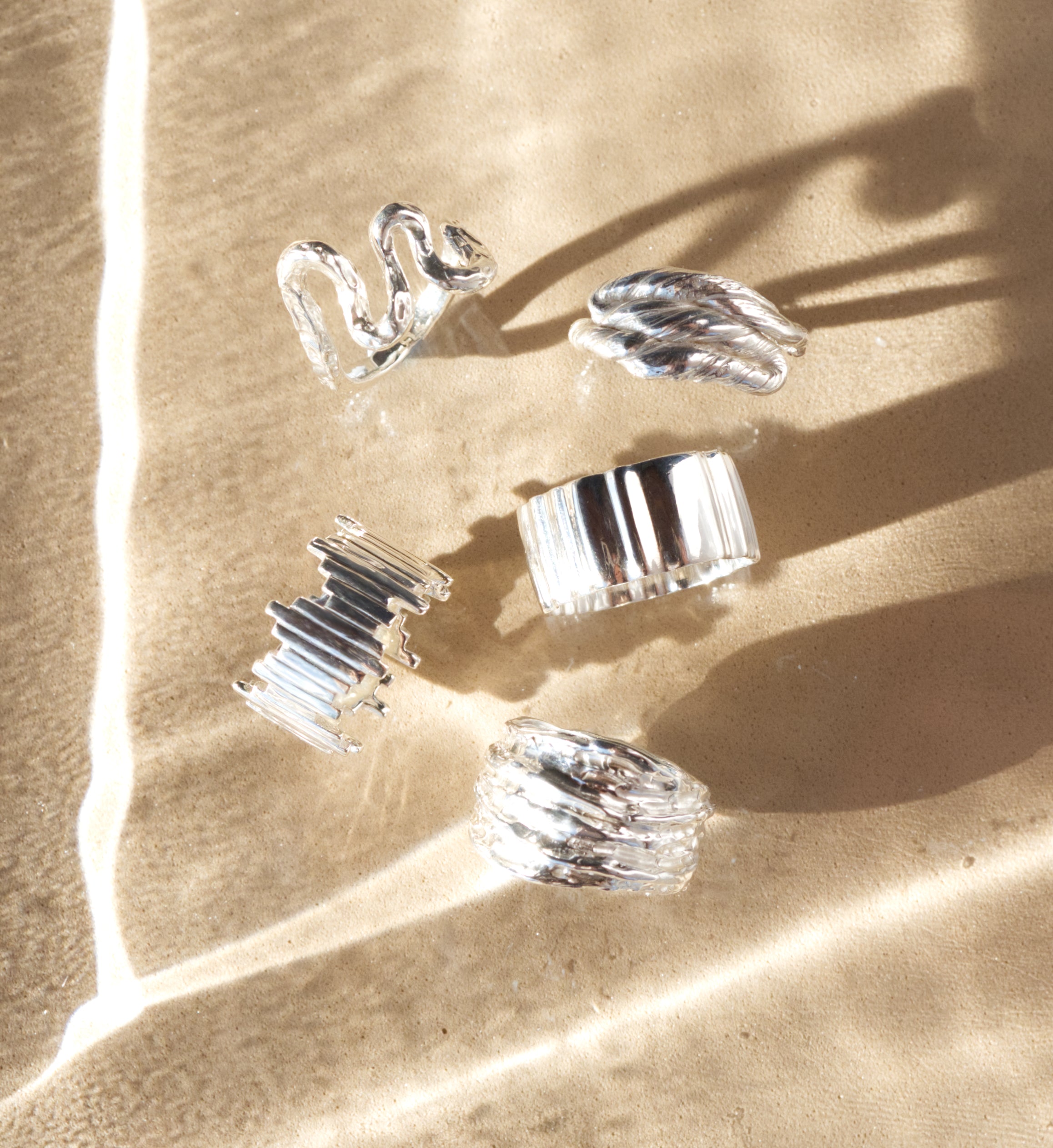 Five silver rings: Flow, Anchor, Wave, Guidance and Canyon, pictured from the top on a sandy textured sunlit background.