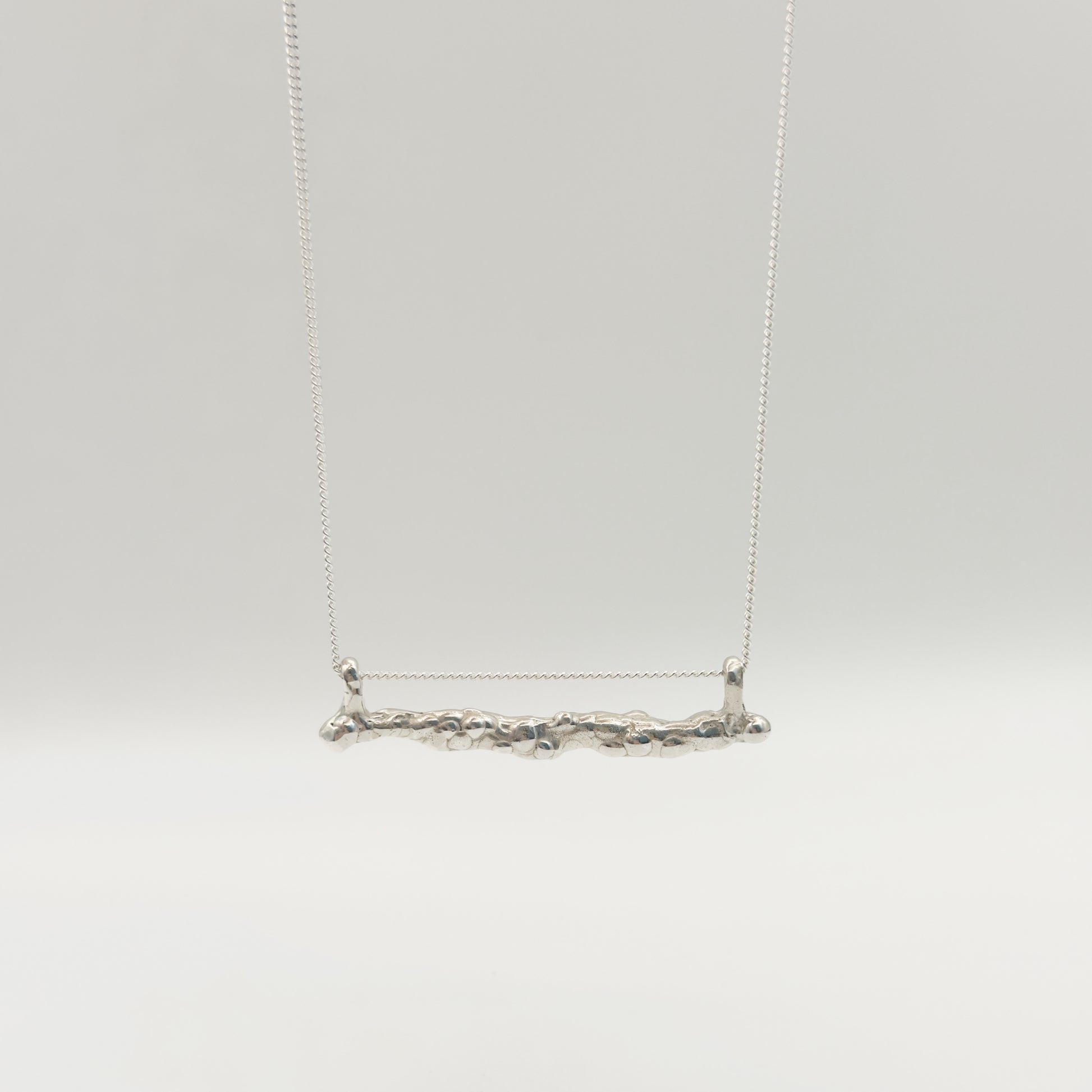 Back view of the Atoll textured silver bar pendant on a chain, pictured on a light plain background.
