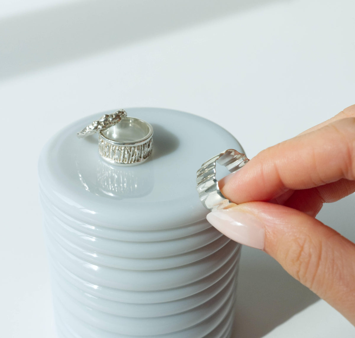 A close-up of a light blue textured round container, with two silver textured rings resting on top - one with a bark-like grooved band and one with a small cluster of beads. A hand's thumb and forefinger hold a third silver ring with a wavy, fluted edge above the stack.