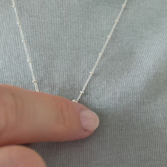 A close up of a hand feeling the texture of the Self-Love necklace. Pictured on a person wearing a light blue top.