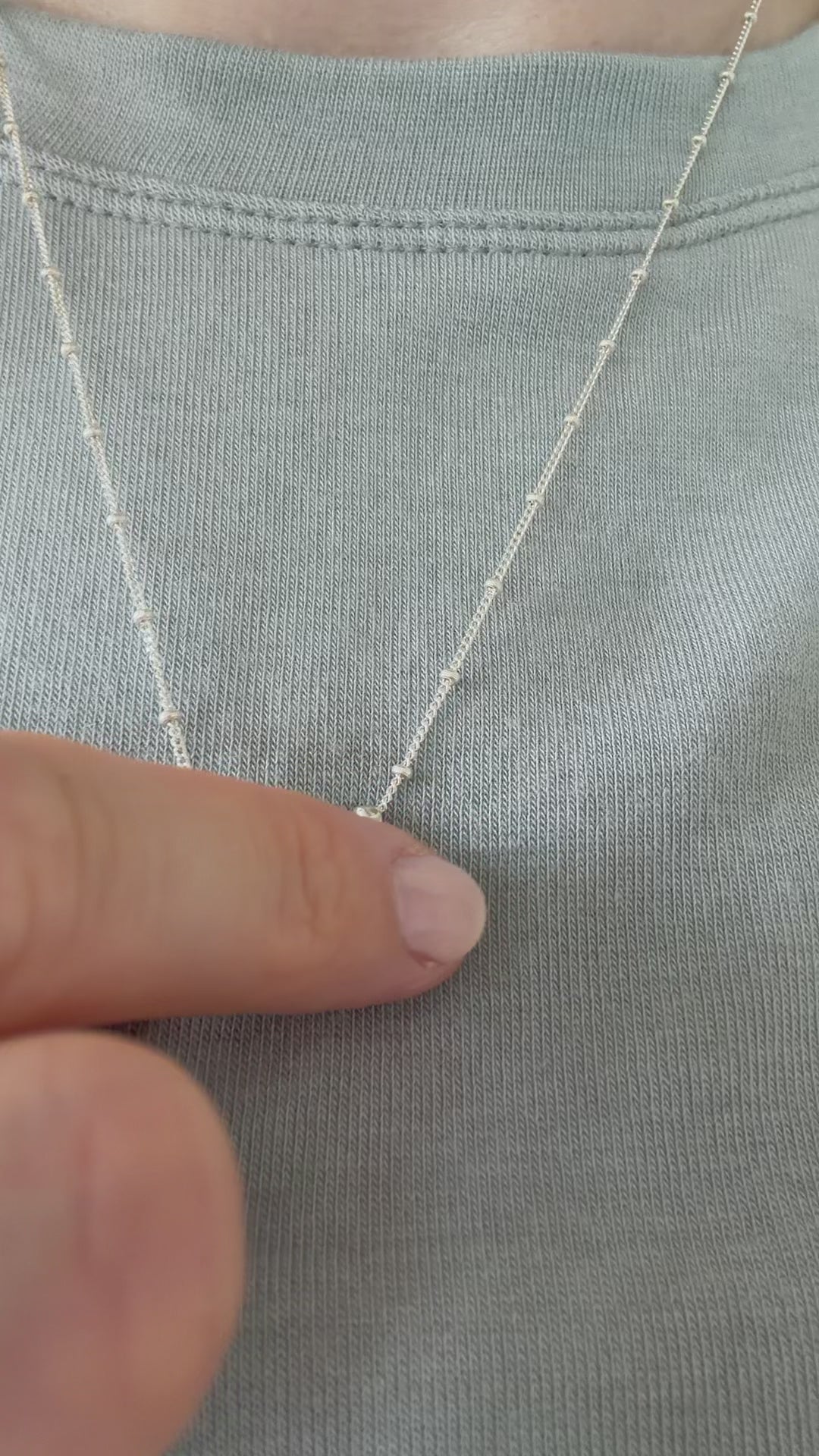 A close up of a hand feeling the texture of the Self-Love necklace. Pictured on a person wearing a light blue top.