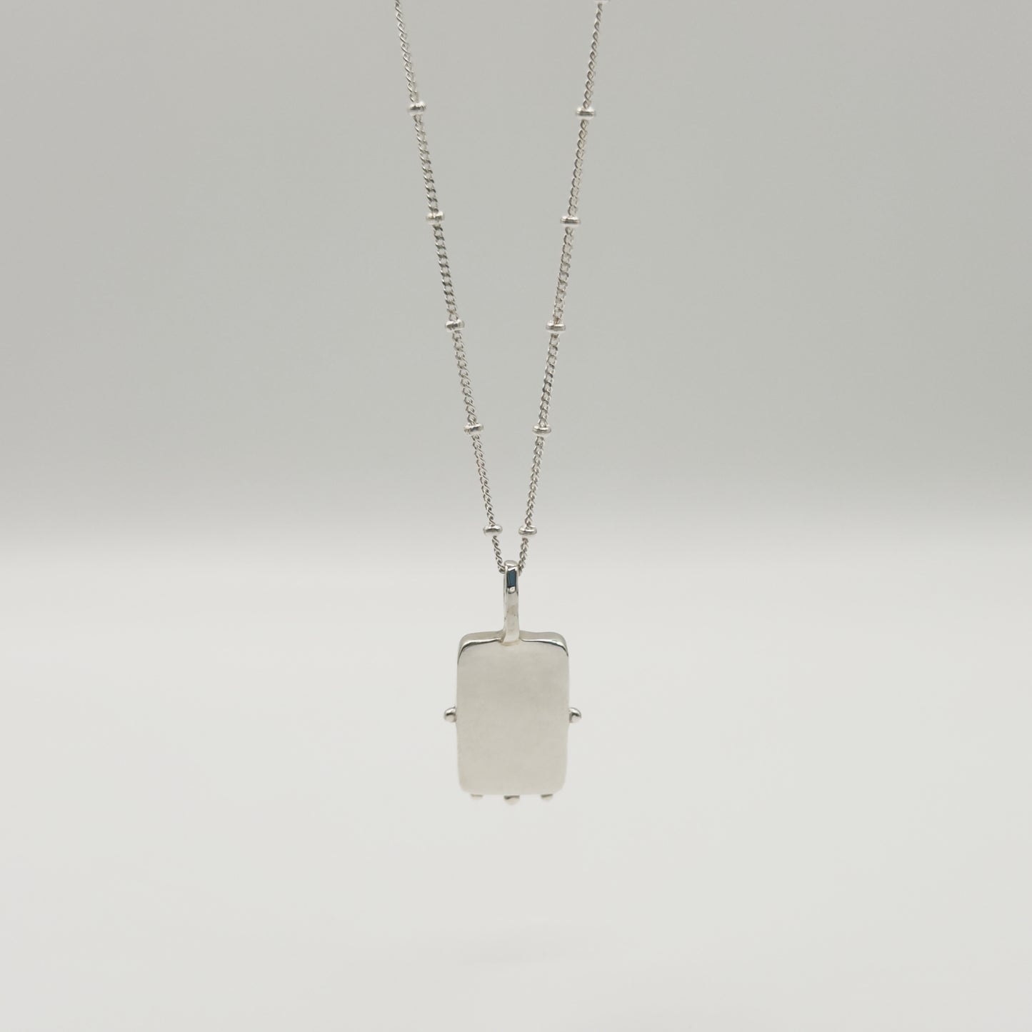 Back view of the Self-Compassion textured silver pendant on a satellite chain, pictured on a light plain background.