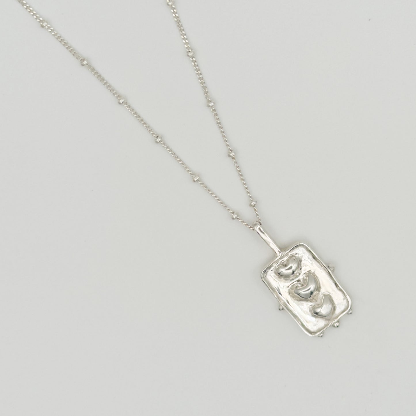 Close-up top view of the Self-Compassion textured silver pendant on a satellite chain, pictured on a light plain background.