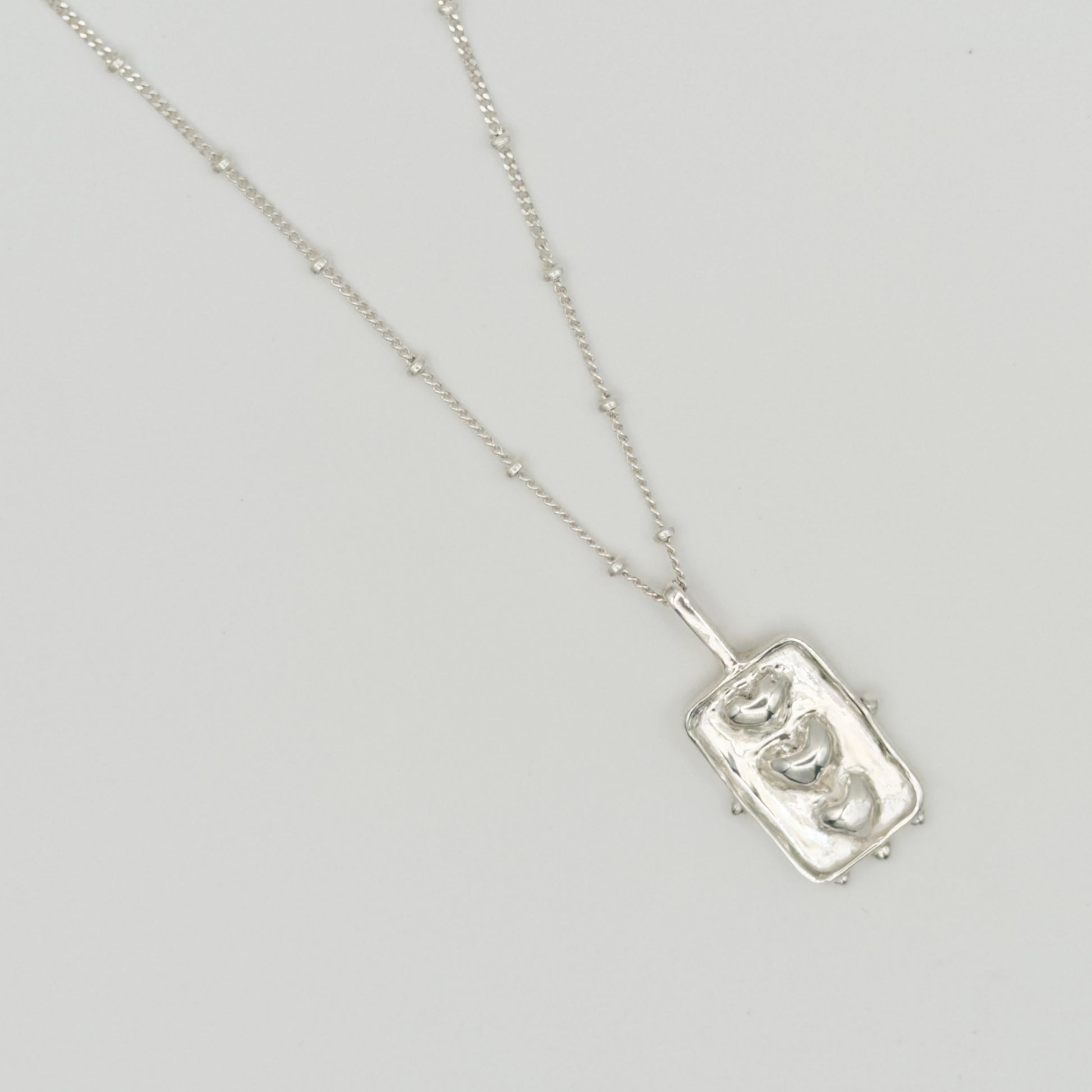 Close-up top view of the Self-Compassion textured silver pendant on a satellite chain, pictured on a light plain background.
