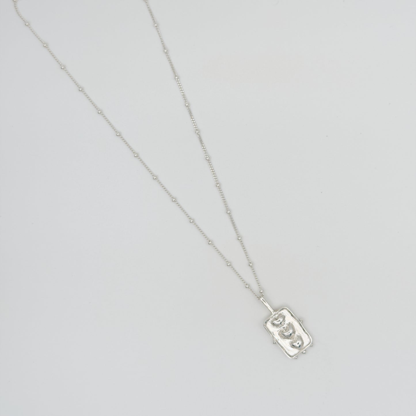 Top view of the Self-Compassion textured silver pendant on a satellite chain, pictured on a light plain background.