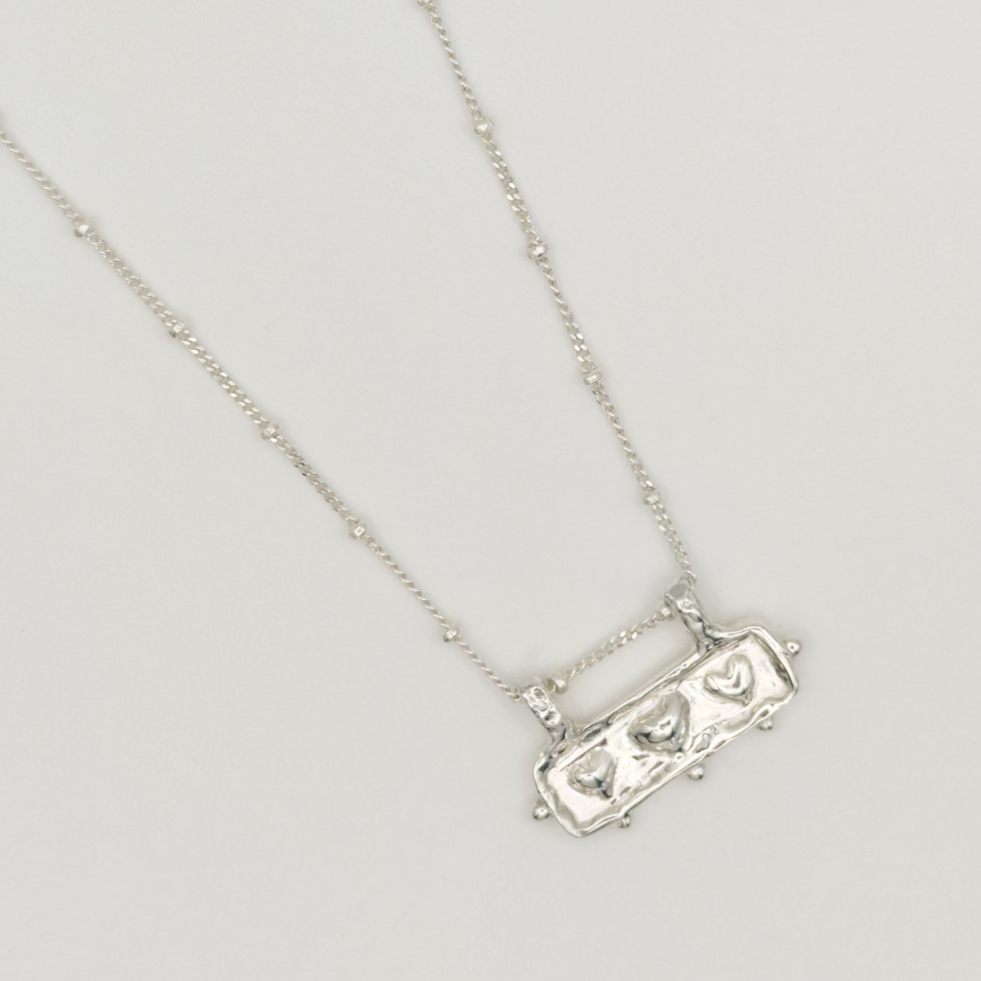 Close-up top view of the Self-Love textured silver pendant on a satellite chain, pictured on a light plain background.