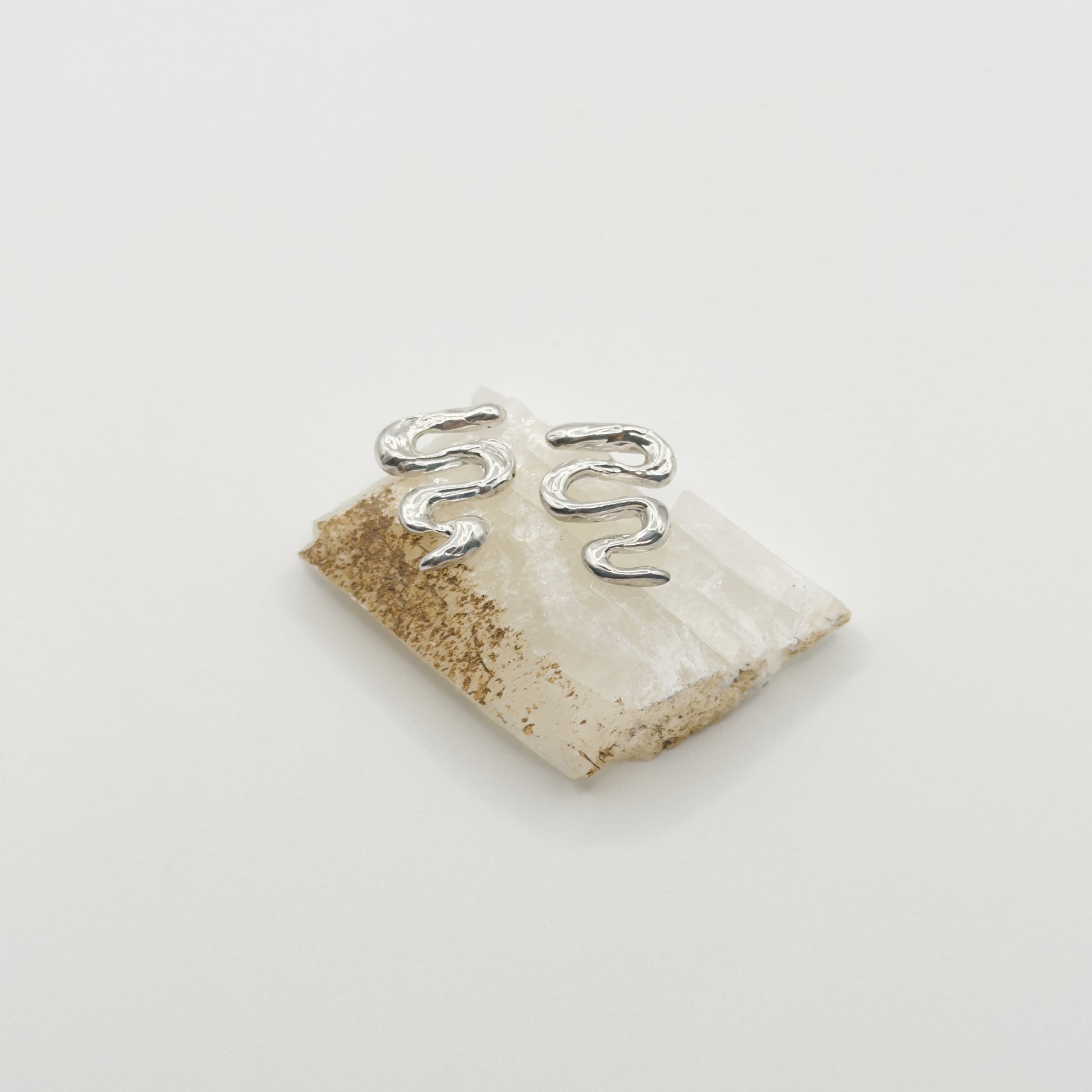 Front view of the Unfold textured silver stud earrings, pictured on a piece of beige rock, on a light plain background.