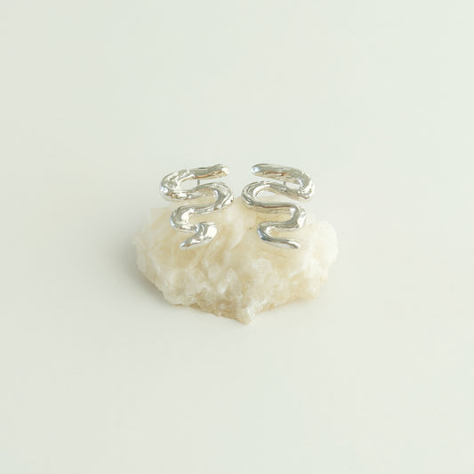 Angled front view of the Unfold textured silver stud earrings, pictured on a piece of beige rock, on a light plain background.